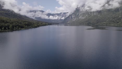 Upward-pullback-drone-video-over-Lake-Bohinj-between-the-mountains,-clear-sky-with-some-clouds