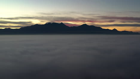 Drone-flying-above-thick-fog-at-sunrise,-silhouetted-mountains-in-distance