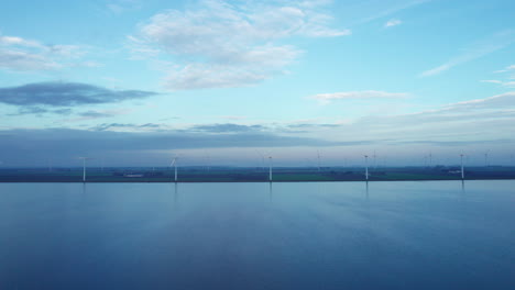 Drone-truck-shot-of-the-lakeshore-covered-in-wind-turbines-on-a-calm-windless-day
