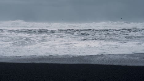 Atemberaubende-Stürmische-Ozeanwellen-Des-Atlantiks-Erreichen-Den-Schwarzen-Sandstrand-Auf-Der-Isländischen-Insel---Vogelschwarm-Fliegt-über-Spritzwasser---Statisch