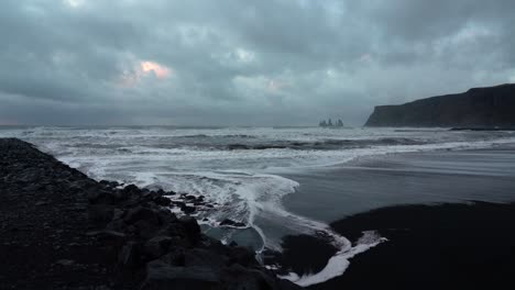 Wide-shot-of-black-beach-with-reaching-waves-in-Vik,Iceland-during-cloudy-day---Famous-Basalt-Sea-Stacks-in-background