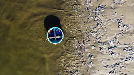 Traditional-Vietnamese-Coracle-boat-and-sandy-beach-full-of-plastic-and-other-dirt,-aerial-top-down-view