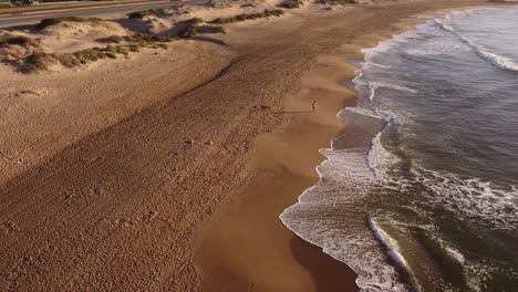 Aerial-top-down-of-Man-walking-on-sandy-beach-beside-Atlantic-Ocean-Waves-in-Sun