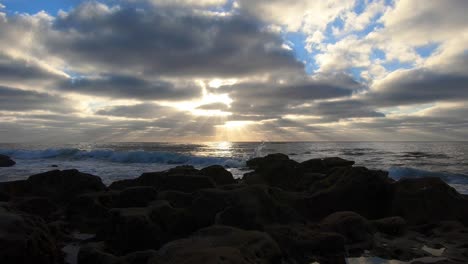 establishing-shot-of-sun-rays-peaking-through-the-clouds-over-the-ocean-on-a-cloudy-day-with-small-waves-rolling-onto-shore-with-rocks-in-the-foreground-and-patches-of-blue-sky