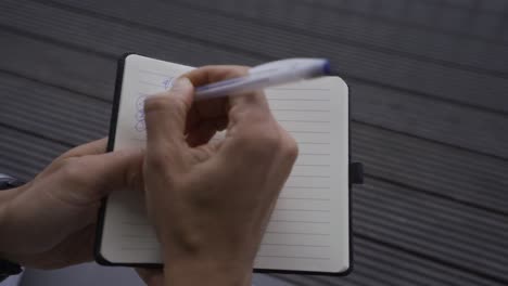 Closeup-of-a-man's-hand-writing-down-the-day's-to-do-list-in-a-small-notebook-on-a-wooden-table