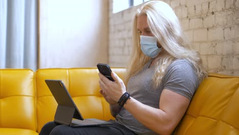 Masked-long-blonde-hair-bearded-man-on-yellow-vegan-leather-vinyl-couch-typing-on-iPad-Pro-Magic-Keyboard-then-picking-up-iPhone-13-Pro-Max-and-texting-messaging-back---in-Cinema-4k-