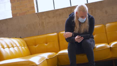 Masked-long-blonde-hair-bearded-man-on-yellow-vegan-leather-vinyl-couch-watching-YouTube-video-or-Netflix-and-Hulu-on-mobile-cellphone-iPhone-13-Pro-Max---in-Cinema-4k-