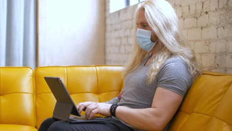 Masked-long-blonde-hair-bearded-caucasian-white-man,-strong-muscular-and-healthy-sitting-on-yellow-vegan-leather-vinyl-couch-typing-on-iPad-Pro-magic-keyboard---in-Cinema-4k-