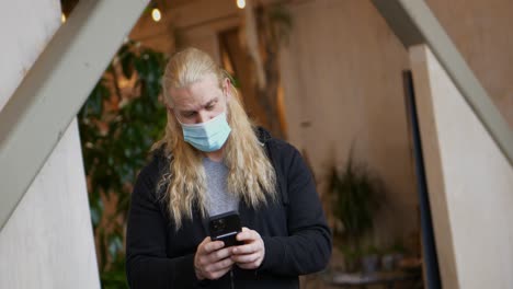 Masked-long-blonde-hair-bearded-man-on-his-cellphone-iPhone-13-Pro-Max-standing-in-public-place-making-facial-gestures-and-playing-with-adjusting-medical-mask---in-Cinema-4k-