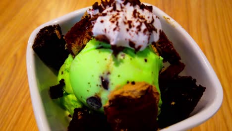 mint-chocolate-chip-ice-cream-double-scoop-in-a-bowl-surrounded-by-chocolate-cake-mini-squares-and-topped-with-whipped-cream-and-rotating-on-a-wooden-surface