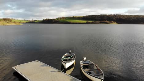Aerial-Low-Flying-Over-Wimbleball-Lake-On-Exmoor-In-Somerset-With-Empty-Boats-Beside-Pier