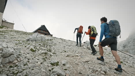 Hikers-with-big-backpacks-going-toward-the-mountain-cottage-climbing-up-on-rocks-and-using-hiking-poles