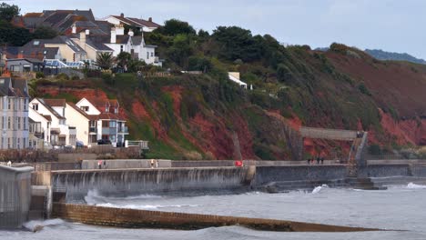 Waves-crash-against-the-sea-wall-at-Dawlish-in-Devon,-UK-as-a-GWR-express-train-passes-along-the-red-cliff-coast