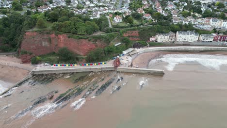 Drone-Aerial-reveal-of-Lea-Mount,-Teignmouth-Hill,-Dawlish-with-beach-huts-and-pier-as-a-train-pulls-out-of-the-tunnel
