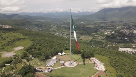 Aerial-drone-rotating-shot-of-large-majestic-patriotic-red,-white-and-green-Mexican-flag-waving-in-wind-along-hilly-terrain-at-daytime