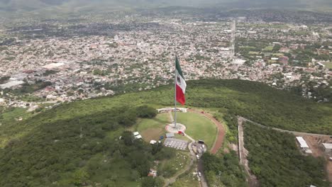 Aerial-drone-fast-moving-shot-of-large-majestic-red,-white-and-green-Mexican-flag-waving-in-wind-along-hilly-terrain-at-daytime