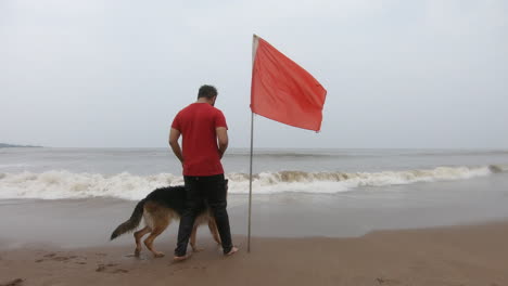 Deutscher-Schäferhund-Am-Strand-Mit-Seinem-Besitzer-In-Der-Nähe-Einer-Roten-Fahne-|-Menschliche-Und-Tierische-Freundschaft-|-Hochalarmflagge-Am-Strand