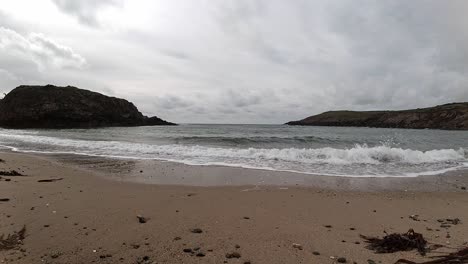 Slow-motion-tide-crashing-on-sandy-Welsh-beach-with-island-mountains-across-horizon