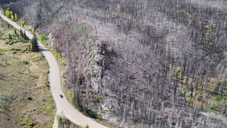 Aerial-view-of-a-truck-driving-down-a-dirt-road-next-to-a-completely-burned-forest-at-the-base-of-a-mountain-in-Colorado