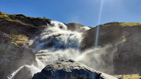 Looking-up-at-beautiful-Norwegian-clean-mountain-waterfall-in-Vikafjell-Voss-Norway---Summer-sun-with-sunbeams-and-blue-sky-background-and-water-splashing-all-over---Static-handheld