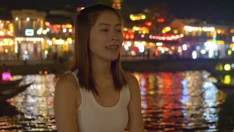 Medium-shot-of-Asian-girl-at-Hoi-An-ancient-town-riverfront-at-night---blurry-boats-on-river-and-colorful-lights-in-background