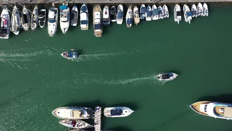 Boat-traffic-in-a-Latchi-port-passing-moored-yachts,-aerial-downwards-view