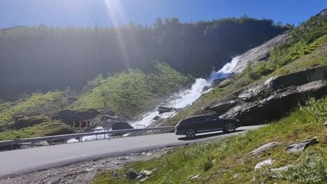 Cars-driving-uphill-Halsabakkane-hill-heading-towards-Vikafjell-mountain-in-Norway-with-beautiful-Sendefossen-waterfall-in-background---Handheld-static-clip-with-sunrays-entering-top-frame