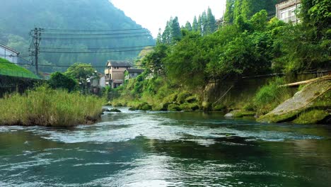 Drone-flight-over-pristine-river-in-peaceful-Japanese-mountainside,-lush-vegetation-surrounding-quaint-village