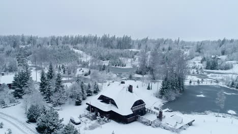 Aerial-over-bright-white-snow-covered-winter-landscape---smallholding-in-rural-woodland-area-next-to-frozen-lake