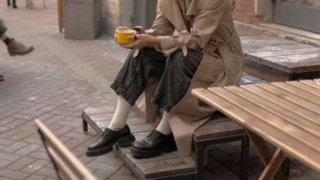Fashionable-woman-is-sitting-on-wooden-stairs-outside-and-drinking-coffee