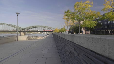 Dolly-forward-shot-of-a-pedestrian-walkway-along-the-Rhein-Treppen,-close-to-a-stone-ledge-with-trees,-bridge,-and-cityscape-in-the-background,-slow-motion,-Cologne-Germany
