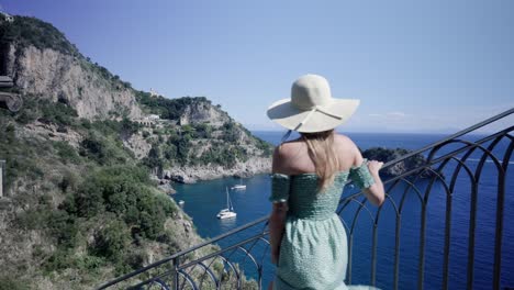 Atractiva-Mujer-Caucásica-Vestida-De-Verano-Con-Sombrero,-Bajando-Las-Escaleras-Con-Impresionantes-Vistas-Costeras-En-Un-Agradable-Día-Soleado-En-Italia
