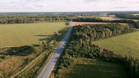 Ascending-drone-flight-over-a-country-road-in-Estonia,-Europe-following-a-lonely-car-driving-along-the-empty-street-between-fields-and-little-forests-in-summer-2022---bird-view-shot-like-advertising
