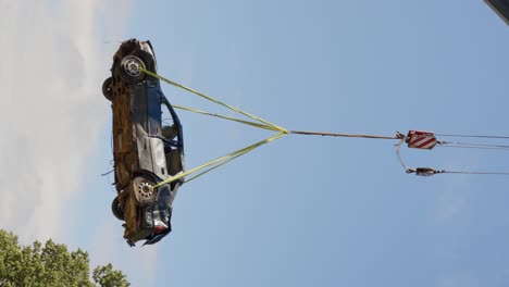 Vertical-video-of-car-wreck-being-lifted-for-recycling-against-blue-sky