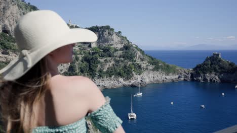 A-Lovely-Caucasian-Woman-Wearing-Off-Shoulder-Summer-Beach-Dress-And-Hat,-Standing-On-Balcony-Watching-The-Scenic-Ocean-View-With-Yachts-Mooring-Offshore-In-An-Island-Resort-In-Italy