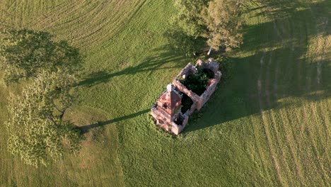 Fast-flyover-lost-place-church-ruin-without-roof-in-Estonia,-Europe---aerial-drone-flight-over-orange-sandstone-building-that-looks-like-a-tiny-orthodox-or-catholic-church-on-a-green-meadow,-2022