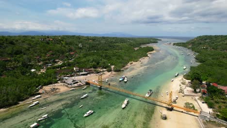 aerial-zoom-out-of-yellow-bridge-over-channel-between-Nusa-Ceningan-and-Lembongan-island-at-low-tide-with-boats-anchored-on-tropical-turquoise-water-in-Bali