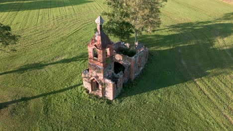 Aerial-drone-flight-over-an-lost-place-church-ruin-without-roof-in-Estonia,-Europe---orange-sandstone-building-that-looks-like-a-tiny-orthodox-or-catholic-church-on-a-green-meadow---bird-view-2022