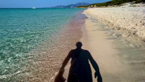 Silhouette-shadow-of-man-taking-off-hat-while-walking-on-sandy-beach-along-turquoise-sea-water-shoreline