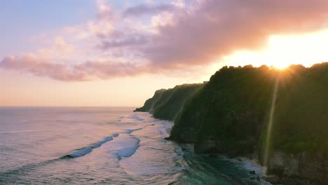 aerial-of-waves-crashing-onto-uluwatu-coastline-with-sun-flare-over-cliffs-at-sunset-in-Bali-Indonesia