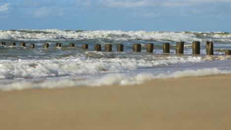 Stormy-waves-breaking-against-old-wooden-pier-on-the-beach,-white-sand-coast,-sunny-day,-Baltic-sea,-distant-low-angle-medium-shot