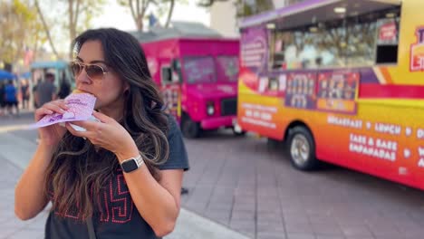 Hispanic-woman-in-the-foreground-eating-food-from-a-food-truck---blurred-background-with-several-food-trucks-and-people-at-a-festival