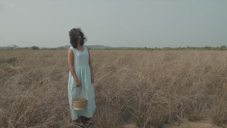 Static-slow-motion-shot-of-a-young-pretty-indian-woman-in-a-light-blue-dress-and-a-wood-basket-in-her-right-hand-standing-on-a-dry-farmer-field-stroking-her-hair-as-the-wind-blows-past-her