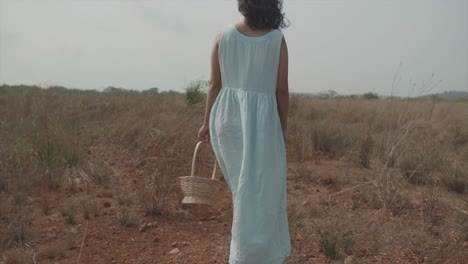 Cinematic-slow-motion-dolly-shot-of-a-young-indian-woman-in-light-blue-dress,-black-hair-and-a-wood-basket-in-her-hands-walking-straight-on-a-dry-and-dusty-field
