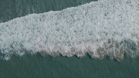 Turquoise-foaming-ocean-breaking-against-sandy-shore-aerial-top-down-view-above-surf
