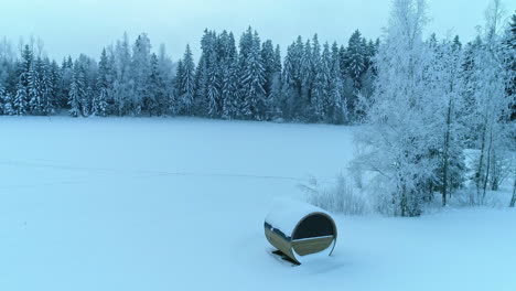 Aerial-pullback-Wooden-barrel-sauna-on-Snow-covered-meadow,-Frost-forest-in-Background---Orbiting