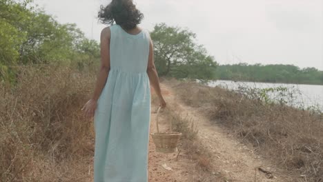 Back-shot-of-a-woman-in-a-blue-dress-walking-in-close-to-the-river-with-a-basket-in-her-hand
