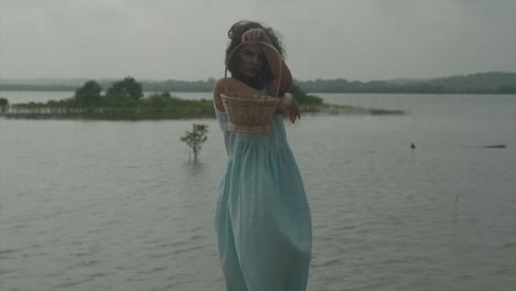 Static-slow-motion-shot-of-a-young-pretty-woman-in-light-blue-dress-and-black-hair-standing-in-front-of-a-lake-with-an-island-of-plants-and-holding-a-wood-basket-in-front-of-her