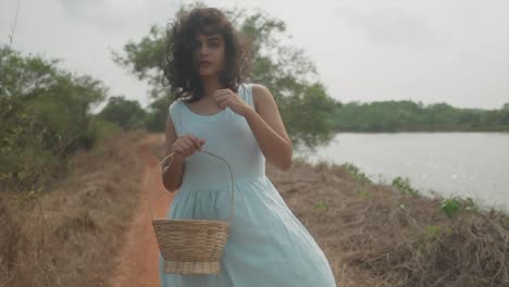 Slow-motion-handheld-dolly-shot-of-an-indian-model-with-turquoise-dress-and-a-wooden-basket-in-her-hand-standing-next-to-a-lake-in-nature-and-looking-into-the-camera