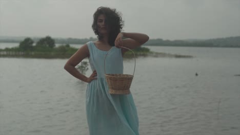 Slow-motion-static-shot-of-a-charming-young-woman-in-light-blue-dress-with-black-hair-standing-by-the-lake-with-a-wood-basket-in-her-hand-in-windy-weather